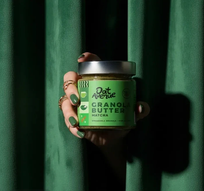 A hand holding a jar of Matcha granola butter through a green velvet curtain